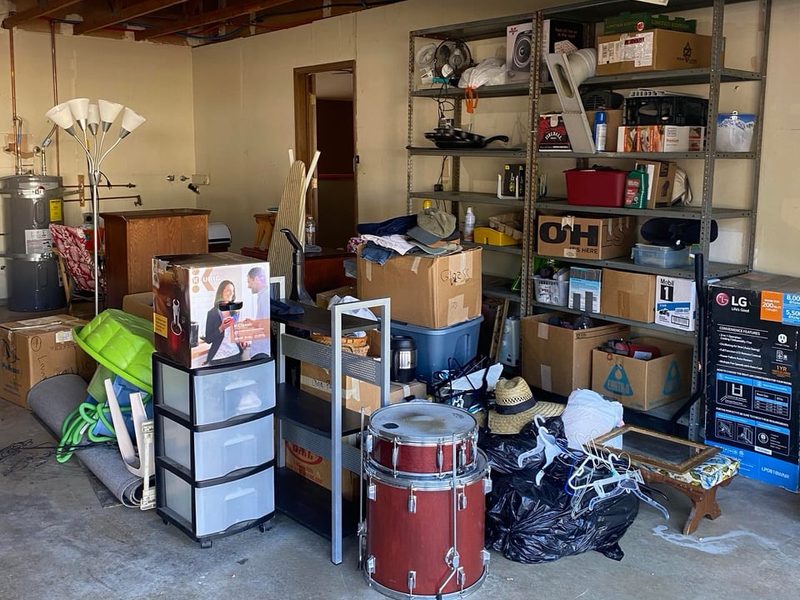 Garage crowded with boxes, a drum kit, a floor lamp and bags of junk