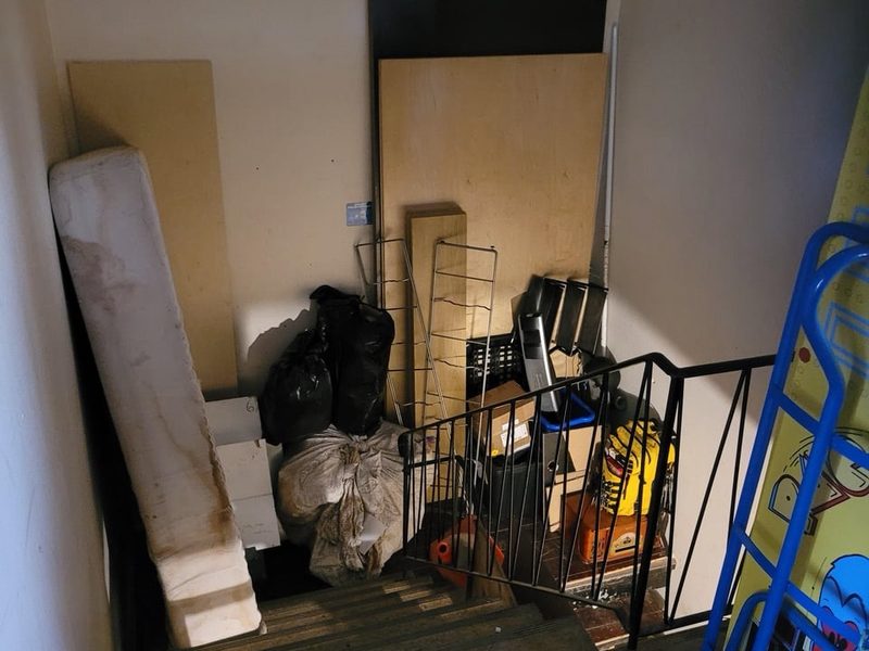 Stairwell landing blocked by an old mattress, boards, bags and a hand truck